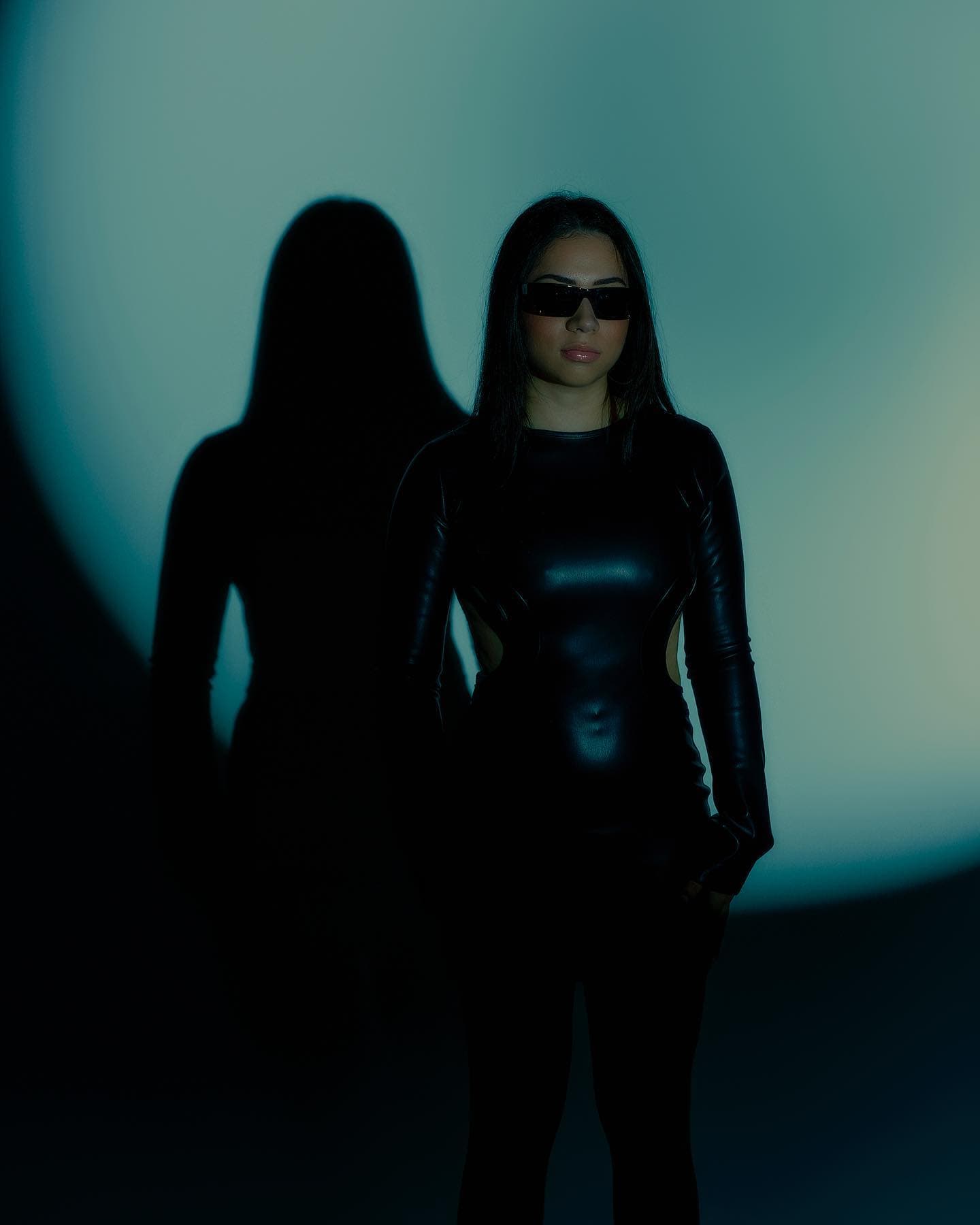 A woman in black standing beside her shadow in a curved field of cyan light